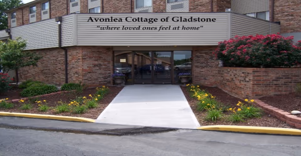 Entrance to Avonlea Cottage of Gladstone, a brick building with a covered walkway and glass double doors. There are flower beds with yellow flowers and green shrubs on either side of the concrete path leading to the entrance.