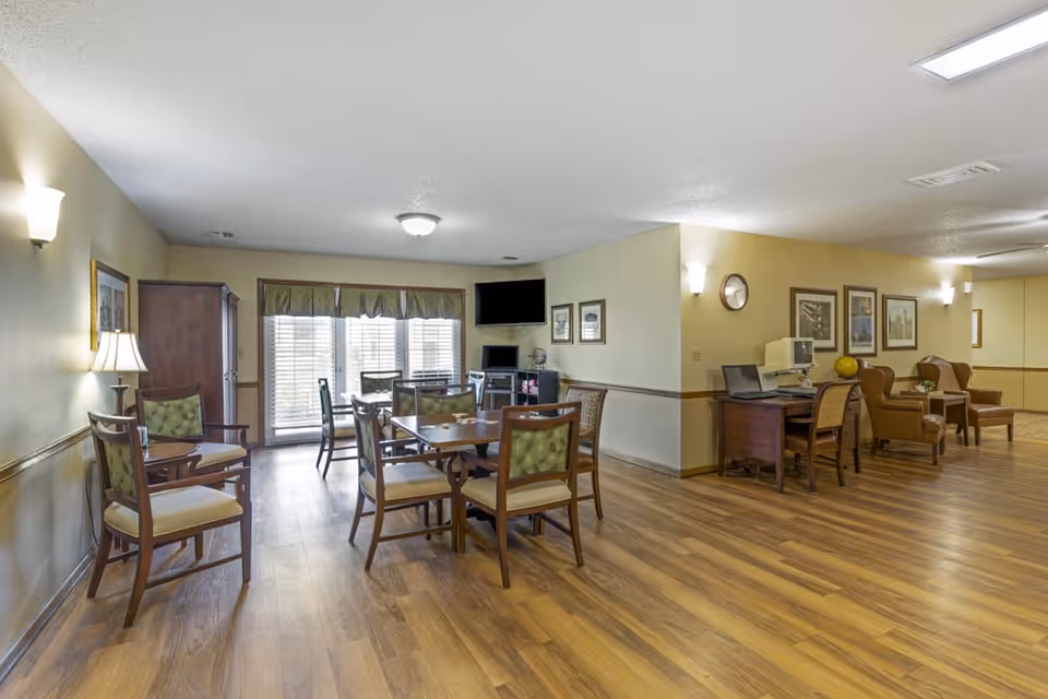 A spacious common area in a senior living facility with wooden flooring, multiple tables and chairs arranged for seating, a TV mounted on the wall, framed pictures, a desk with a computer, and comfortable armchairs along the wall. Large windows with blinds allow natural light to enter the room.
