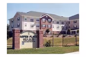 Exterior view of Aspen Place, a multi-story senior living facility with beige siding and white trim. The building has multiple windows and a pitched roof. In front of the building is a brick sign with the name Aspen Place and the address, surrounded by a black metal fence and green grass.