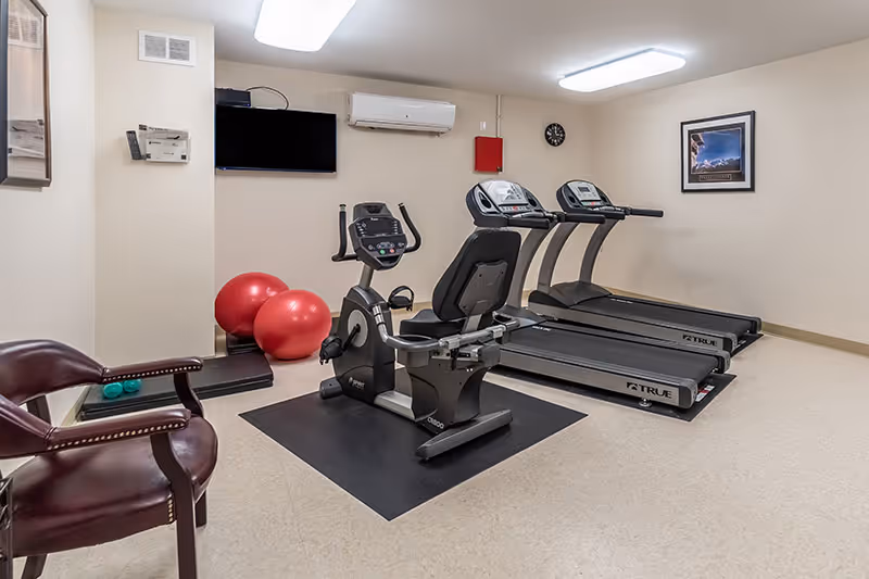 Small fitness room with two treadmills, a recumbent exercise bike, red stability balls, a wall-mounted TV, and chairs.