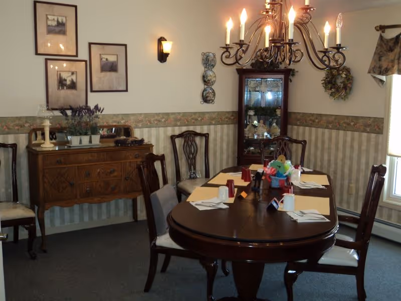 A traditional dining room with a dark wooden oval table set for six people. The table has placemats, napkins, salt and pepper shakers, and a colorful centerpiece. Surrounding the table are six wooden chairs with cushioned seats. The room features wallpaper with a floral border, a wooden sideboard with decorative items and framed pictures above it, a glass-fronted cabinet with collectibles, a wall-mounted light fixture, and a chandelier with lit candles. A window with a floral valance allows natural light into the room.