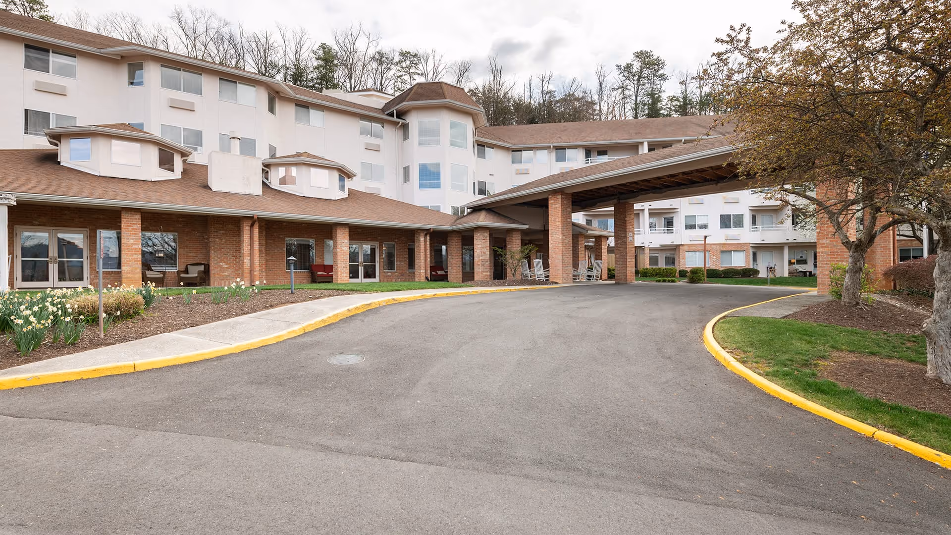 Front exterior view of Holiday Elm Park Estates, a multi-story senior living facility with a covered entrance driveway, brick and white facade, landscaped flower beds, and trees.