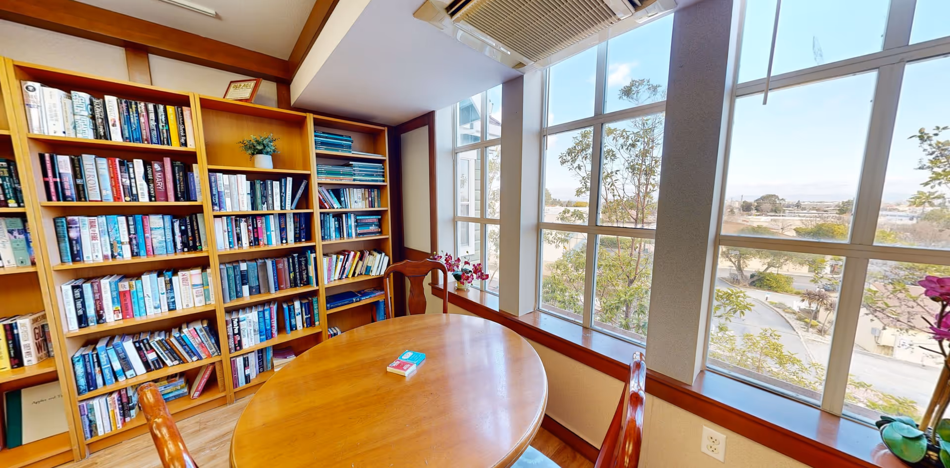 A cozy reading room with wooden bookshelves filled with books, a round wooden table with two wooden chairs, and large windows letting in natural light and showing a view of trees and a parking area outside.