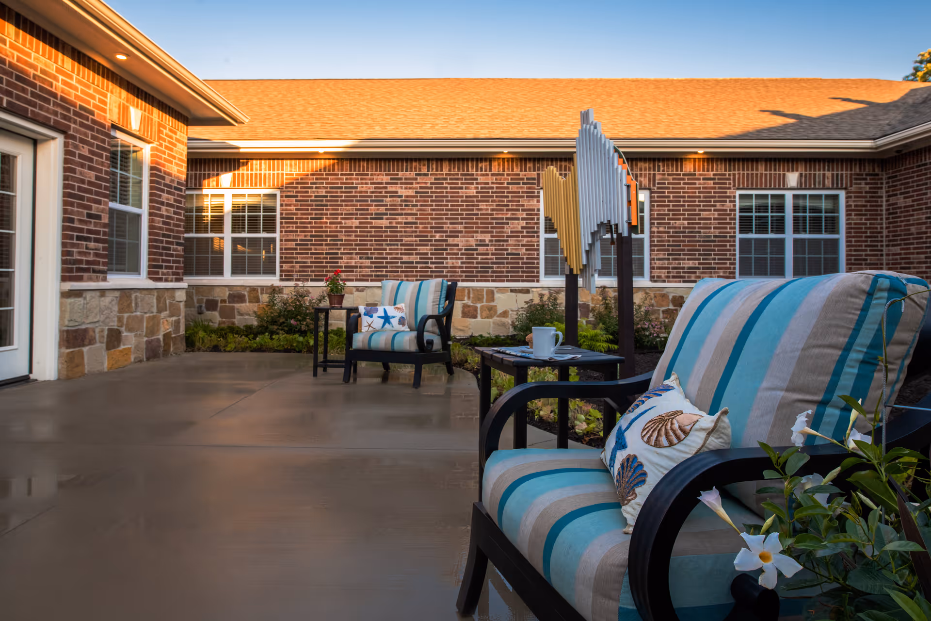 Outdoor patio area at Silverado Overland Park Memory Care Community featuring cushioned chairs with striped upholstery and seashell-themed pillows, small tables with a coffee cup and plant, surrounded by brick and stone building walls and windows.