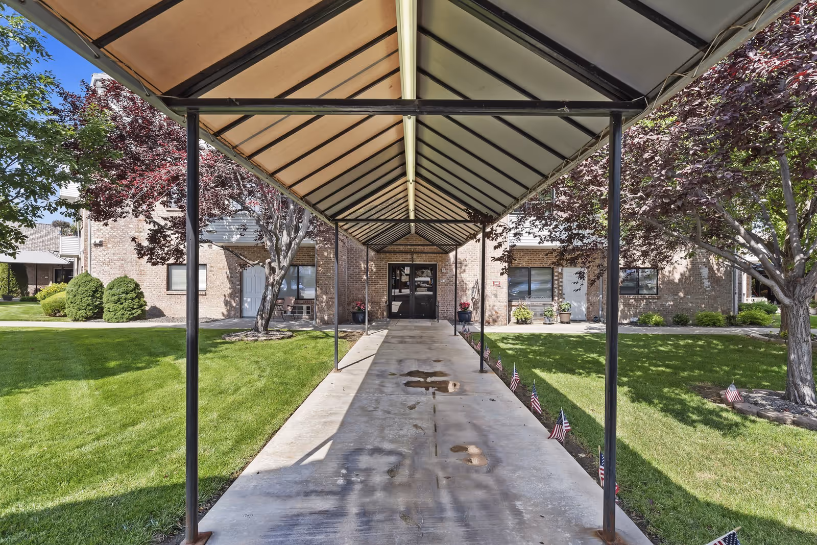 Covered walkway with a metal frame and beige canopy leading to the entrance of a brick building, surrounded by green grass, trees, and small American flags along the walkway edges.