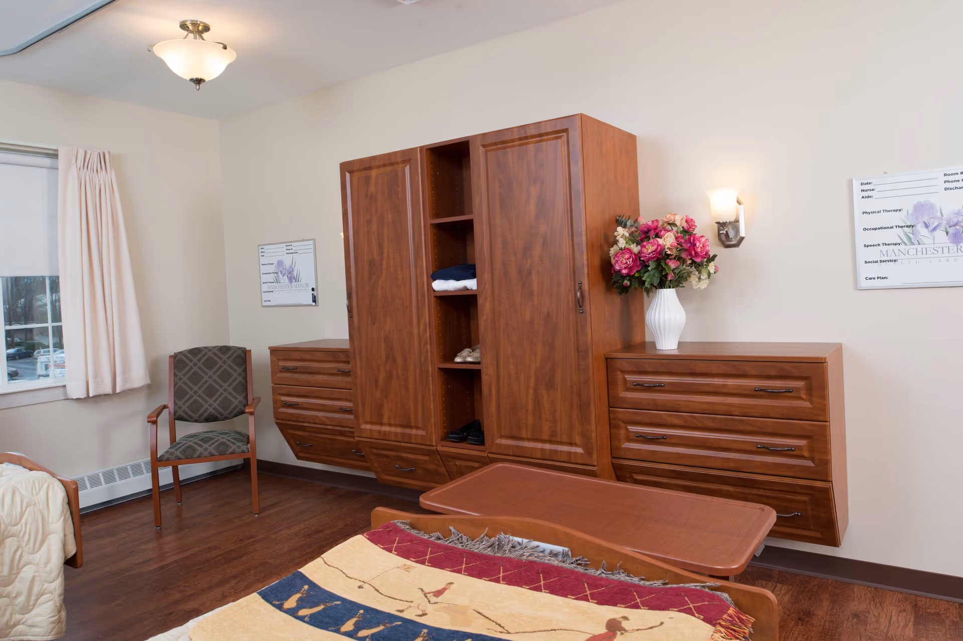 A senior bedroom featuring a wooden wardrobe and matching dressers, a bed with a patterned blanket, and a chair by the window.