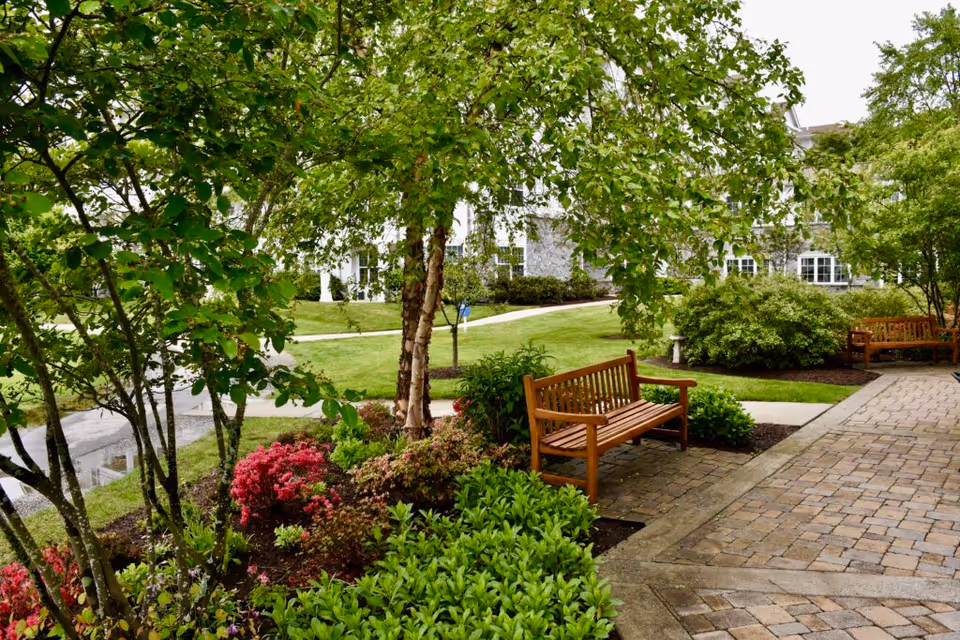 A peaceful outdoor garden area at StoneRidge Senior Living featuring green trees, colorful flowering bushes, two wooden benches, and a paved walkway with a building visible in the background.