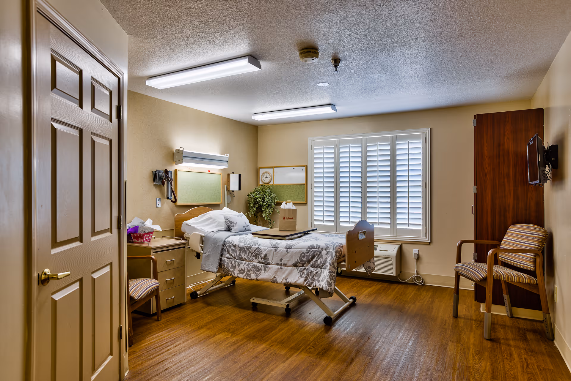 A senior living facility room with a hospital-style adjustable bed covered with a patterned blanket, a bedside table with a chair, a wall-mounted phone, a window with white plantation shutters, a wooden cabinet, a wall-mounted TV, and a striped armchair. The room has wood flooring and beige walls.
