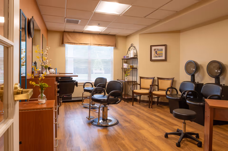 Interior view of a salon area in a senior living facility with wooden flooring, two black salon chairs with footrests, two hair drying stations with black chairs, a small black rolling stool, and a shelving unit with decorative items. There are also two beige chairs with wooden arms against the wall and a window with blinds and a valance letting in natural light.