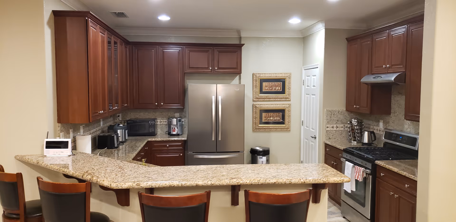 A modern kitchen with dark wooden cabinets, granite countertops, and stainless steel appliances including a refrigerator and stove. There is a breakfast bar with four chairs in the foreground. The kitchen is well-lit with recessed ceiling lights and has decorative framed artwork on the wall near a white door.