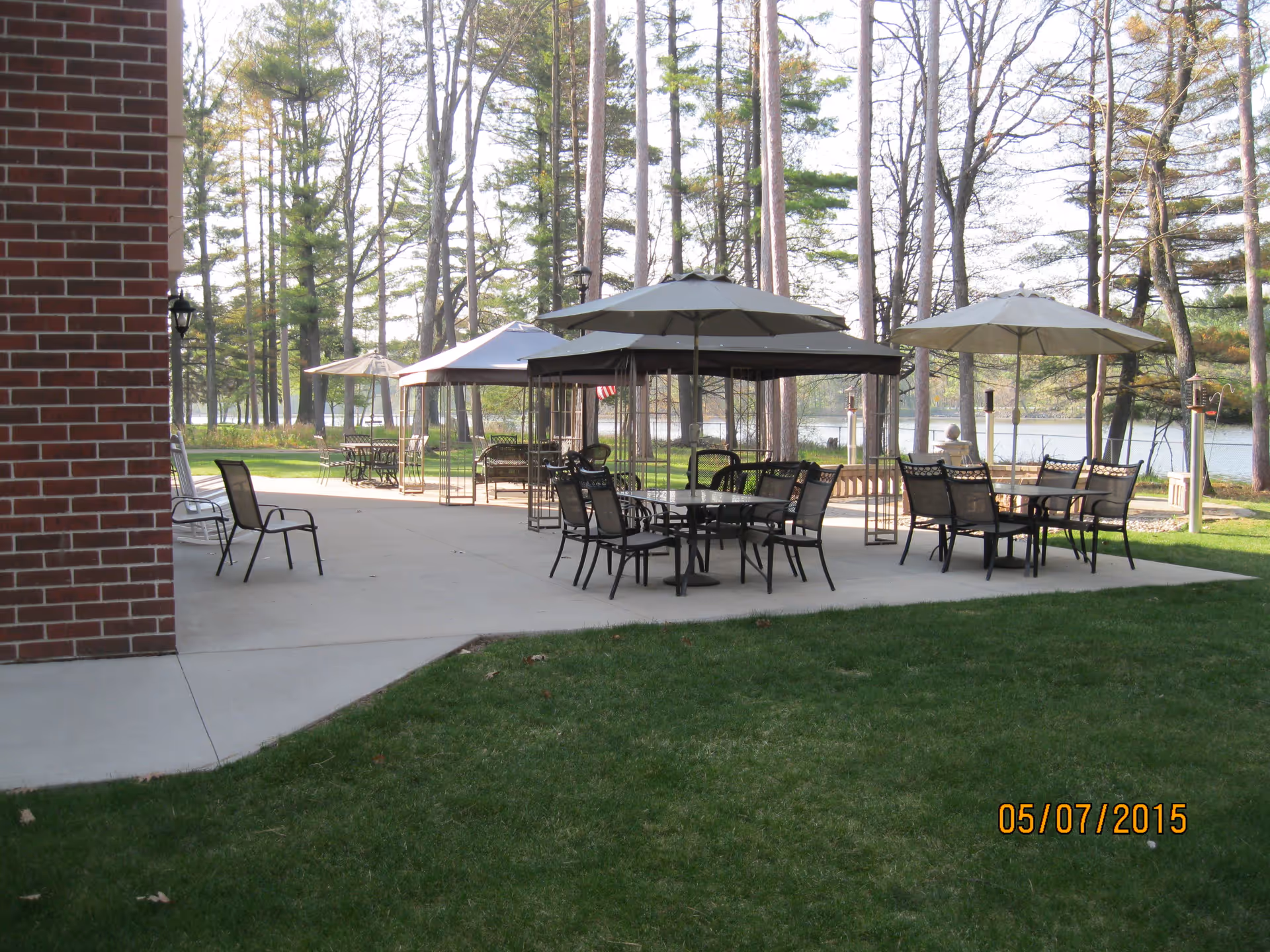 Outdoor patio area with several tables and chairs under large umbrellas and canopies, surrounded by tall trees and overlooking a body of water. A brick building wall is visible on the left side, and green grass is in the foreground.