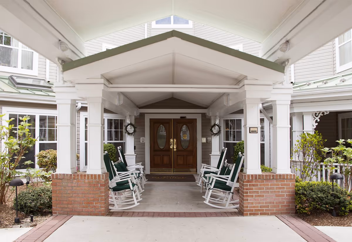 Covered entrance to a senior living facility with a peaked roof supported by white columns on brick bases. There are green cushioned rocking chairs arranged in two rows facing each other along the walkway leading to double wooden doors with oval glass panels. Shrubs and plants are visible on either side of the entrance.