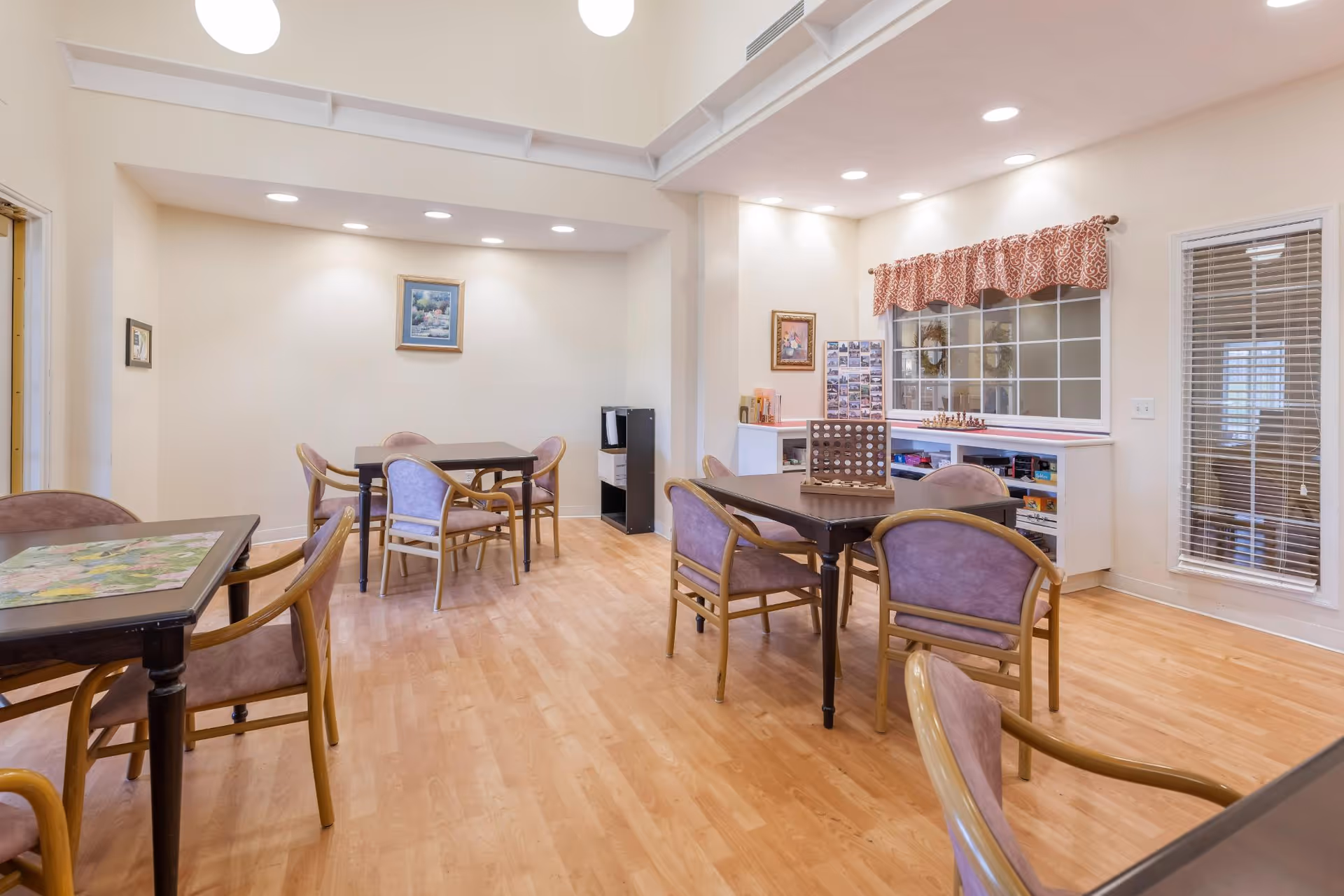 Bright common activity room with several tables and chairs, bookshelves and games along the far wall, and wood flooring.