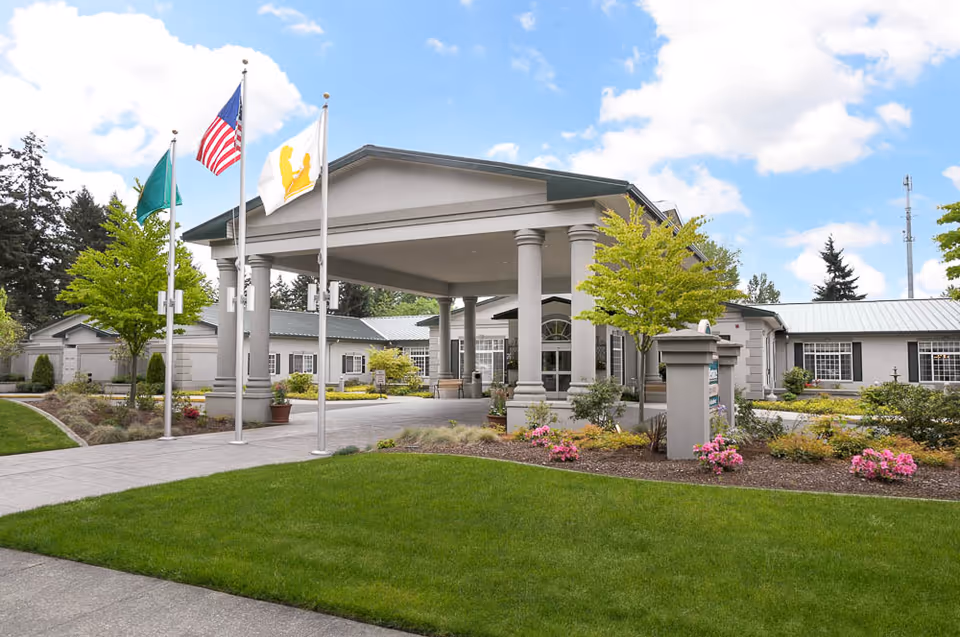 Front exterior view of Garden Terrace Healthcare Center of Federal Way showing a covered entrance with columns, three flagpoles with flags, well-maintained landscaping with green grass, trees, and flowering plants under a partly cloudy sky.