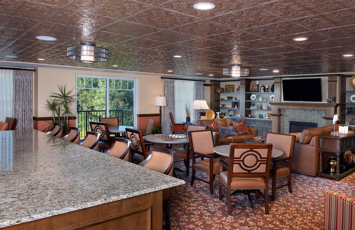 A spacious and well-lit living room area with a granite countertop bar and several bar stools in the foreground. The room features multiple round tables with chairs, a seating area with brown leather sofas and colorful pillows, built-in shelves with decorative items, a fireplace, and a large window with curtains letting in natural light.