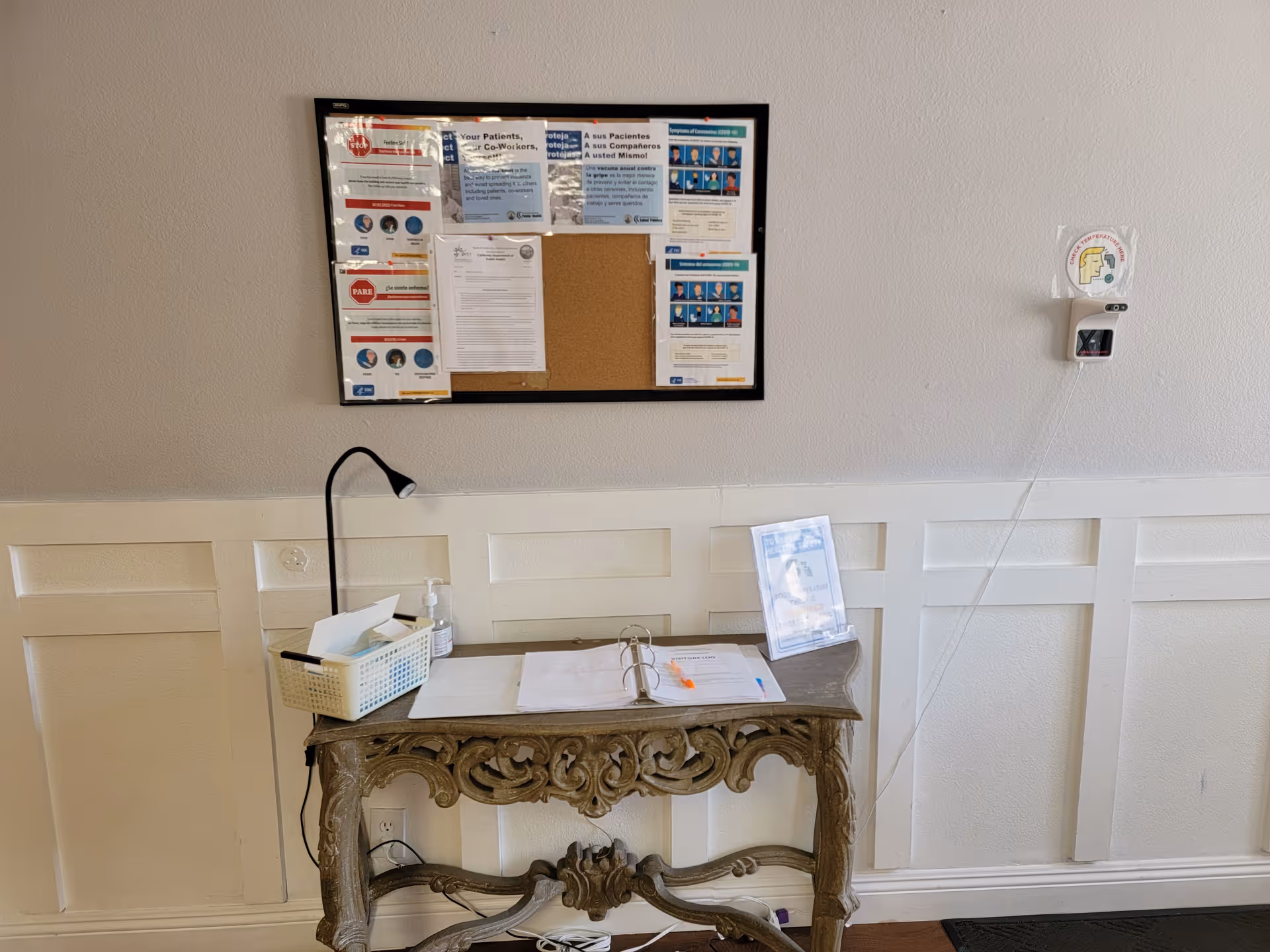 A small ornate wooden table against a white paneled wall with a black desk lamp, a basket with tissues and hand sanitizer, an open binder with papers and a pen, and a clear stand holding a sign. Above the table is a bulletin board with various notices and information sheets. A wall-mounted hand sanitizer dispenser is visible to the right of the table.