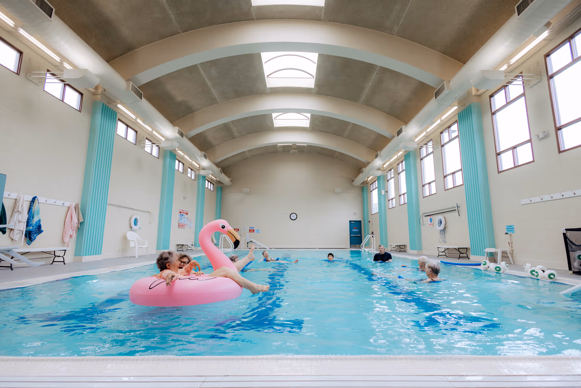 Indoor swimming pool at a senior living facility with residents in the water and a large pink flamingo float under an arched ceiling.
