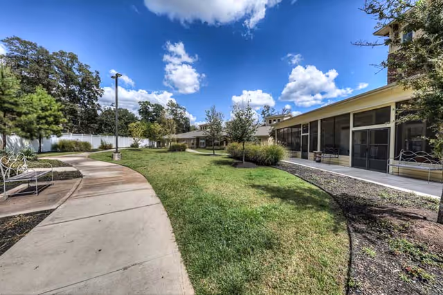 Outdoor view of a senior living facility with a curved concrete walkway, green grass, trees, benches, and a building with large screened windows under a blue sky with scattered clouds.