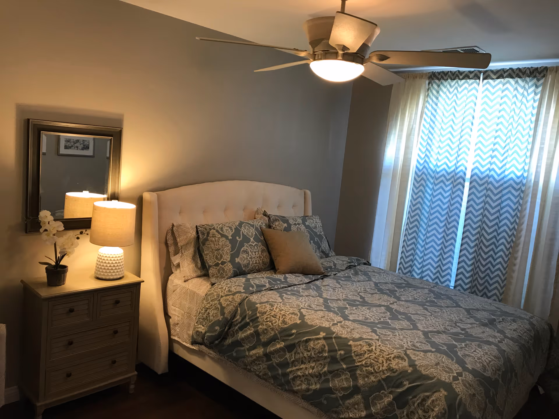A cozy bedroom with a bed featuring a tufted beige headboard and patterned blue and beige bedding. There is a wooden nightstand with drawers on the left side of the bed, holding a white textured lamp and a small potted plant with white flowers. A rectangular mirror hangs on the wall above the nightstand. The room has a ceiling fan with a light fixture and a window covered by sheer curtains with a chevron pattern.