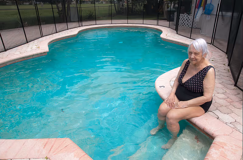 An elderly woman with white hair wearing a black swimsuit with white polka dots sits on the edge of a swimming pool with her feet in the water. The pool area is enclosed with a black safety fence and surrounded by a paved deck.