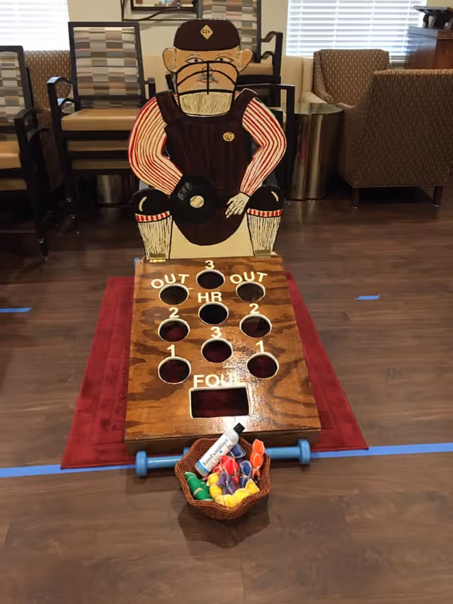 Wooden carnival-style beanbag toss game featuring a painted catcher figure set up on a red rug in a lounge with chairs in the background.
