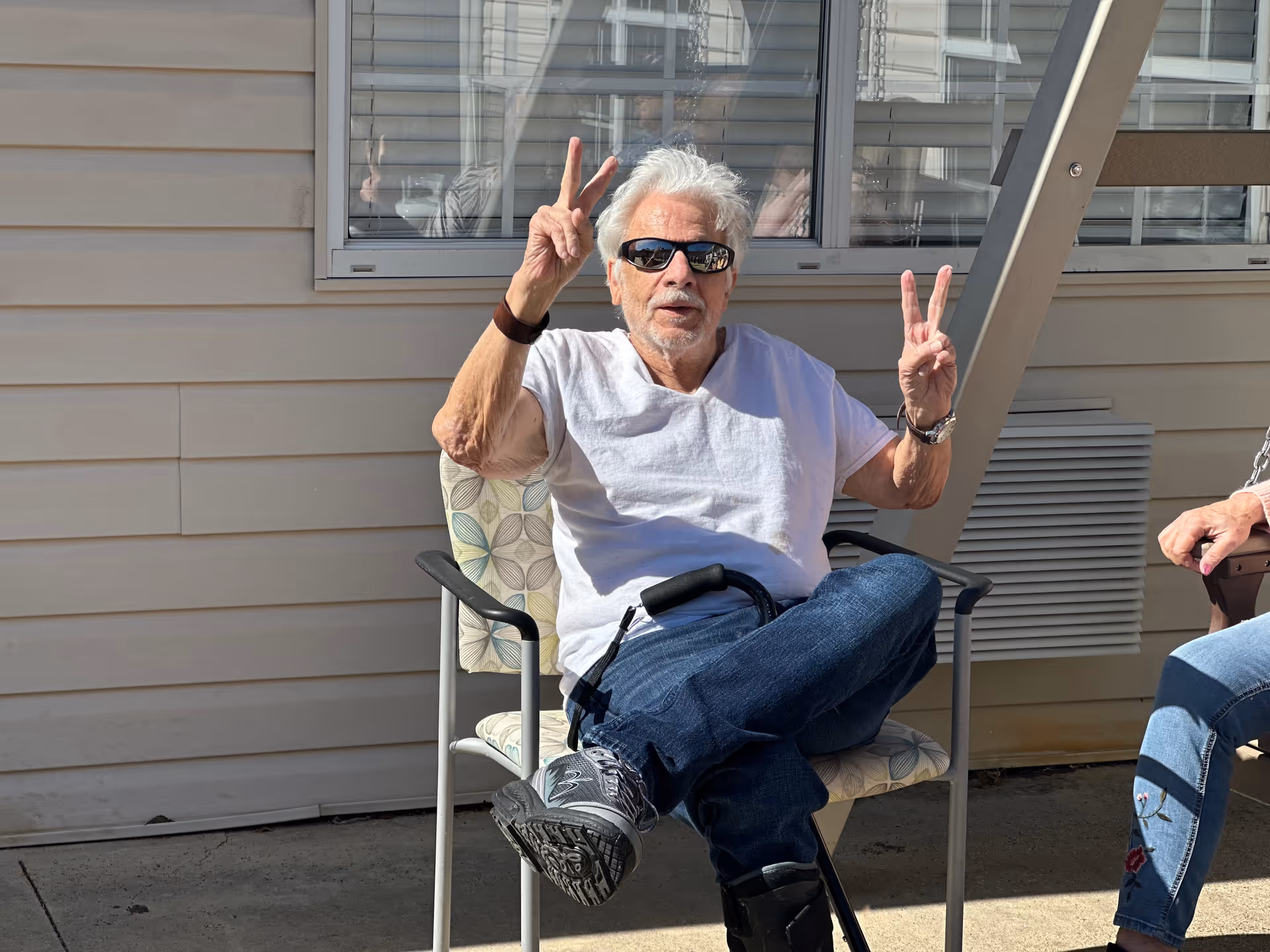 An elderly man wearing sunglasses, a white t-shirt, and jeans is sitting on a patterned chair outdoors in front of a building with beige siding and a window. He is making peace signs with both hands and has a walking cane resting on his lap. Another person is partially visible sitting nearby.