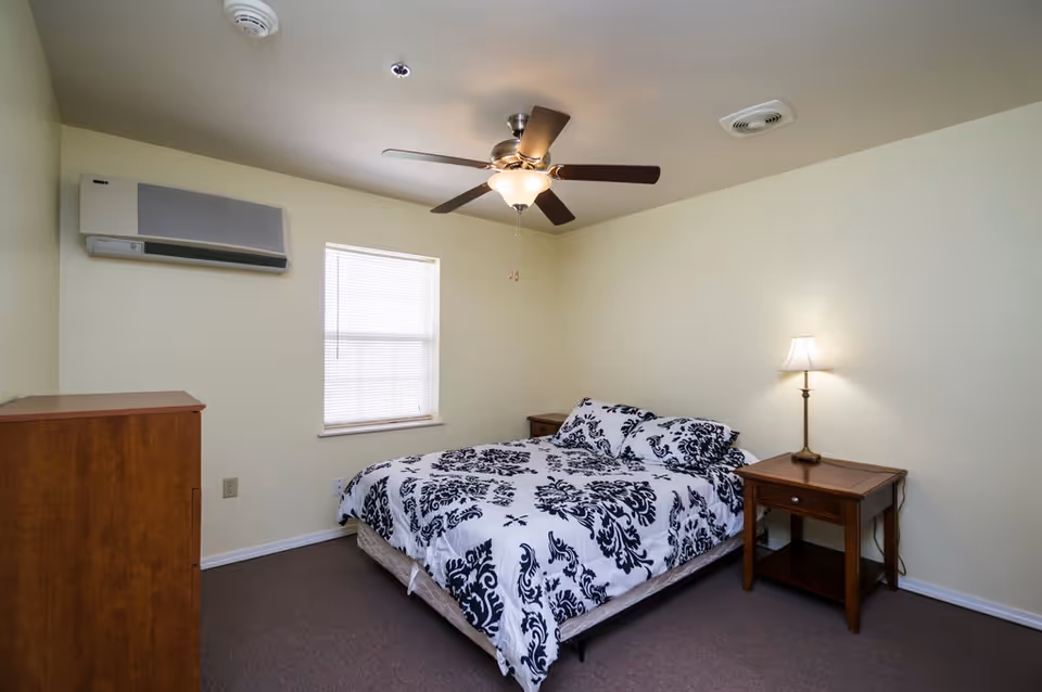 A simple bedroom with a bed covered in a white and black patterned comforter, a wooden nightstand with a lamp, a wooden dresser, a window with blinds, and a ceiling fan with a light.