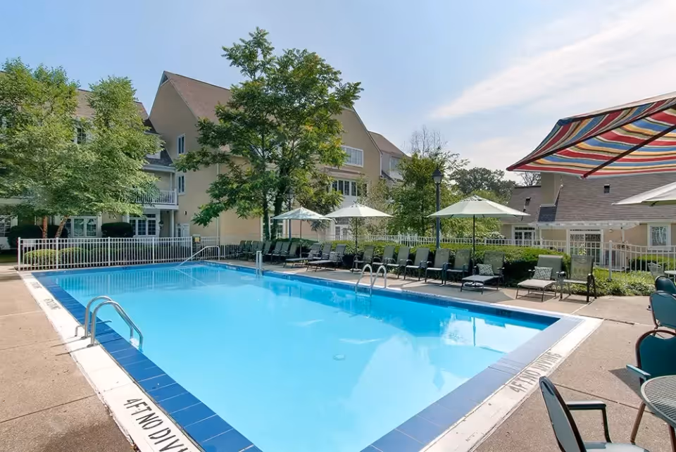 Outdoor swimming pool surrounded by lounge chairs and umbrellas, with a multi-story residential building and trees in the background under a clear sky.