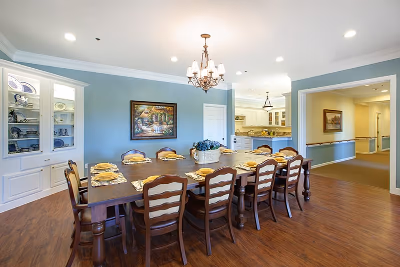 A spacious dining room with a long wooden table set for ten people with yellow plates and placemats. The room has light blue walls, a chandelier hanging from the ceiling, a built-in white cabinet displaying dishes, and a framed painting on the wall. The dining room opens into a kitchen area and a hallway with beige walls and carpet.