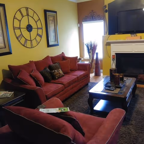 A cozy living room with a red sofa and armchair, a wooden coffee table with decorative items, a large wall clock, framed artwork, a fireplace with a mounted TV above it, and a window letting in natural light.