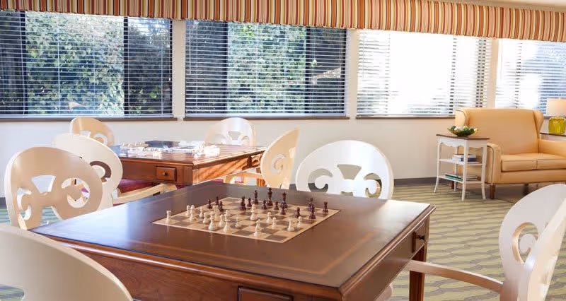 A bright and cozy common area with large windows and striped valances letting in natural light. There are two wooden tables with built-in chess boards and chess pieces set up, surrounded by white chairs with decorative backs. In the background, there is a beige sofa, a side table with a lamp, and a small plant, creating a welcoming and comfortable atmosphere.