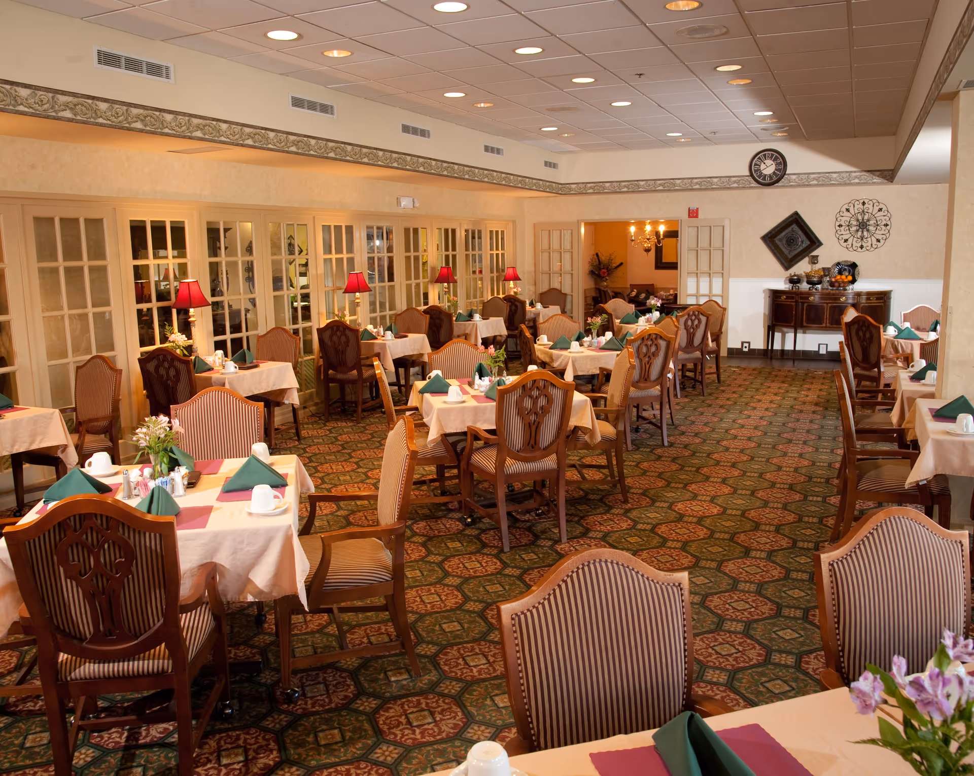 A spacious dining room with multiple tables covered in beige tablecloths, each set with green folded napkins, white cups, and small flower arrangements. The room features patterned carpet, wooden chairs with striped upholstery, red lamps on side tables, and decorative wall art including a clock and ornamental pieces. French doors line one side of the room.