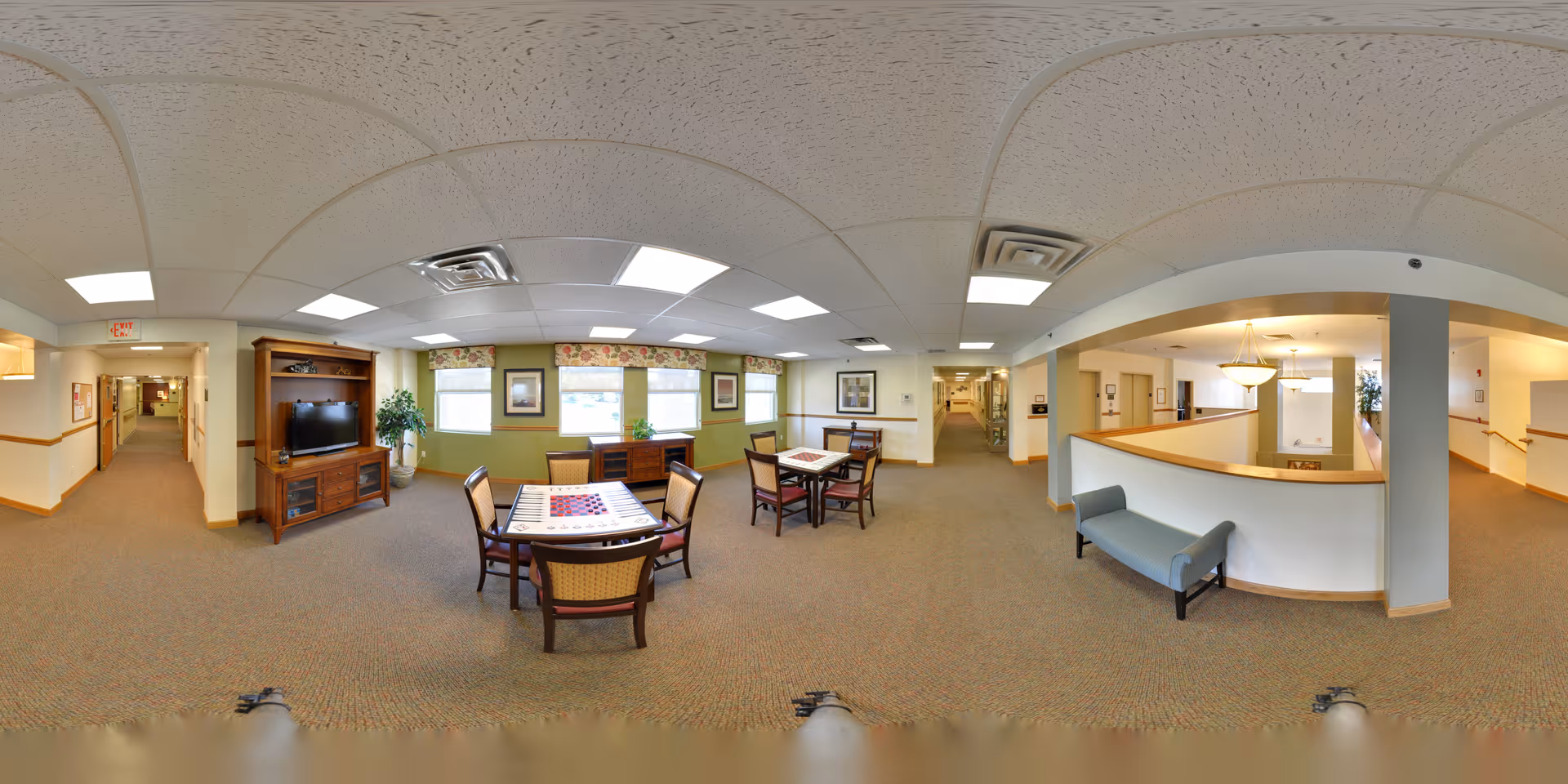A spacious common area in a senior living facility featuring two tables with chairs, a television on a wooden stand, large windows with floral valances, and a hallway leading to other rooms. There is a seating bench near a half-wall with a chandelier above, and the area is well-lit with ceiling lights and neutral-colored walls and carpet.