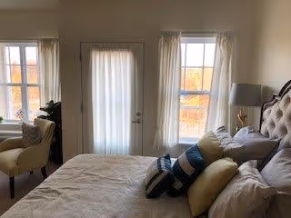 Sunlit bedroom with a tufted bed topped with multiple pillows, an armchair, and a glass door flanked by two windows with sheer curtains.