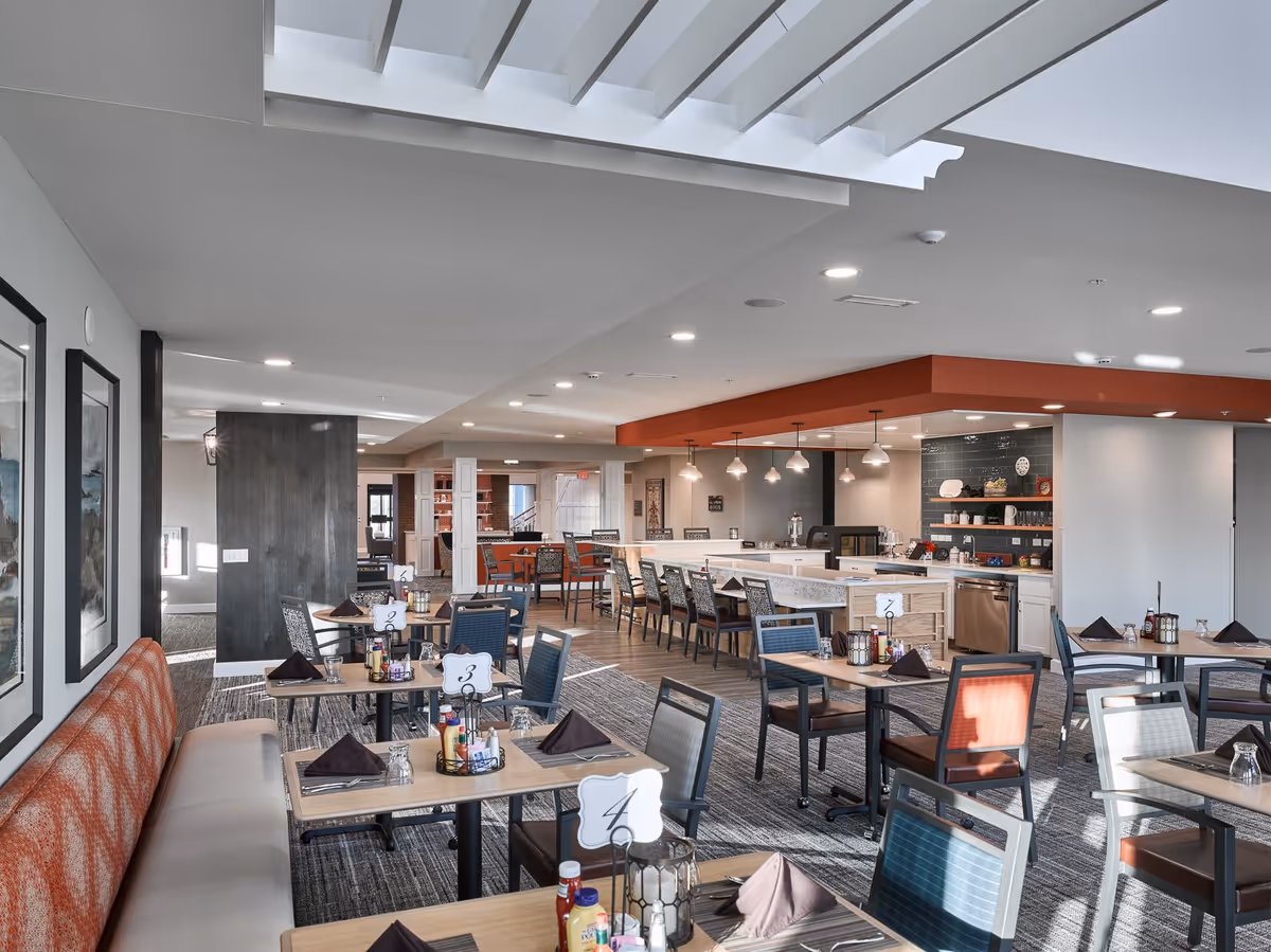 A bright and modern dining room in a senior living facility with multiple tables and chairs arranged neatly. Each table is set with napkins, glasses, and condiments. There is a long counter with bar stools and pendant lights hanging above it. The room features a mix of carpet and wood flooring, with framed artwork on the walls and a large window letting in natural light.