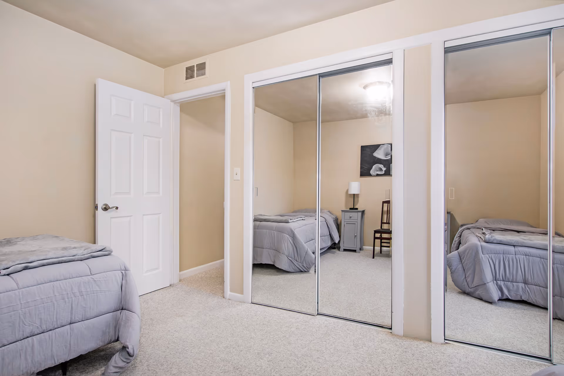 A bedroom with beige walls and carpeted floor featuring a bed with gray bedding on the left side. There is a white door slightly open leading to another room. On the right side, there are large sliding closet doors with mirrors reflecting the bed, a small gray nightstand with a lamp, a wooden chair, and a black and white floral artwork on the wall.