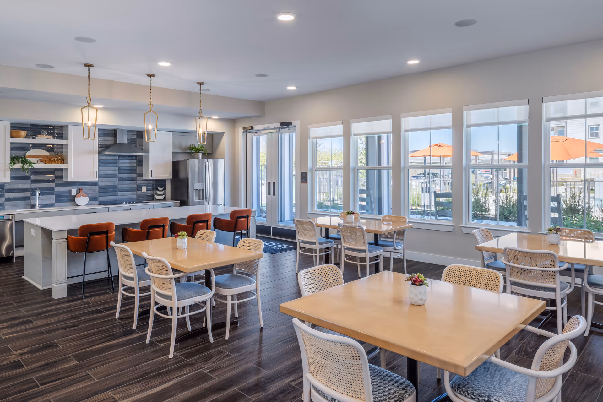 A bright and modern dining area with several light wood tables and white chairs with woven backs. In the background, there is a kitchen area with a large island, orange cushioned bar stools, white cabinets, stainless steel appliances, and pendant lights hanging from the ceiling. Large windows let in natural light and show an outdoor patio with orange umbrellas.