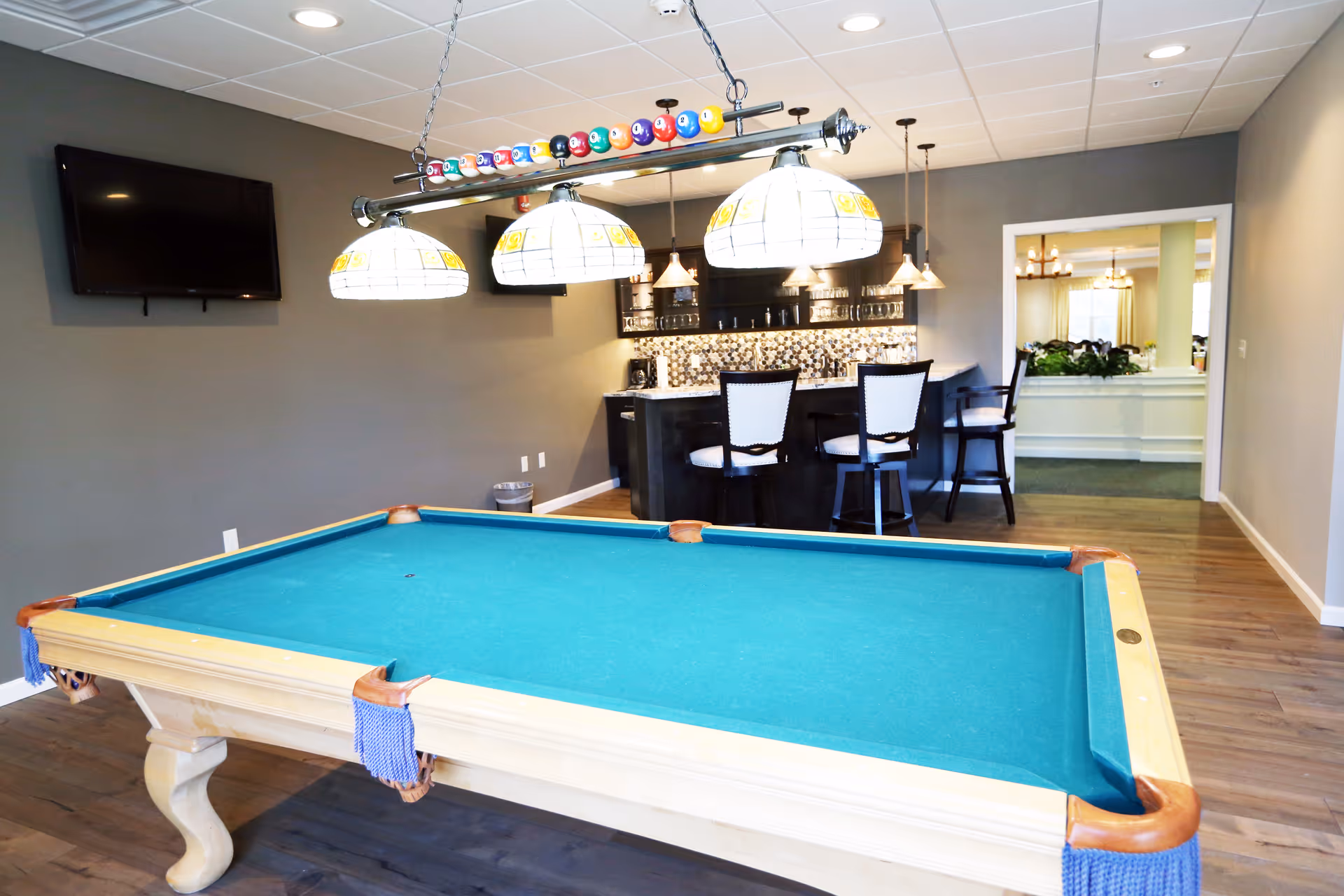 Bright recreation room with a pool table in the foreground, overhead billiard lights, a wall-mounted TV and a small bar with stools in the background.