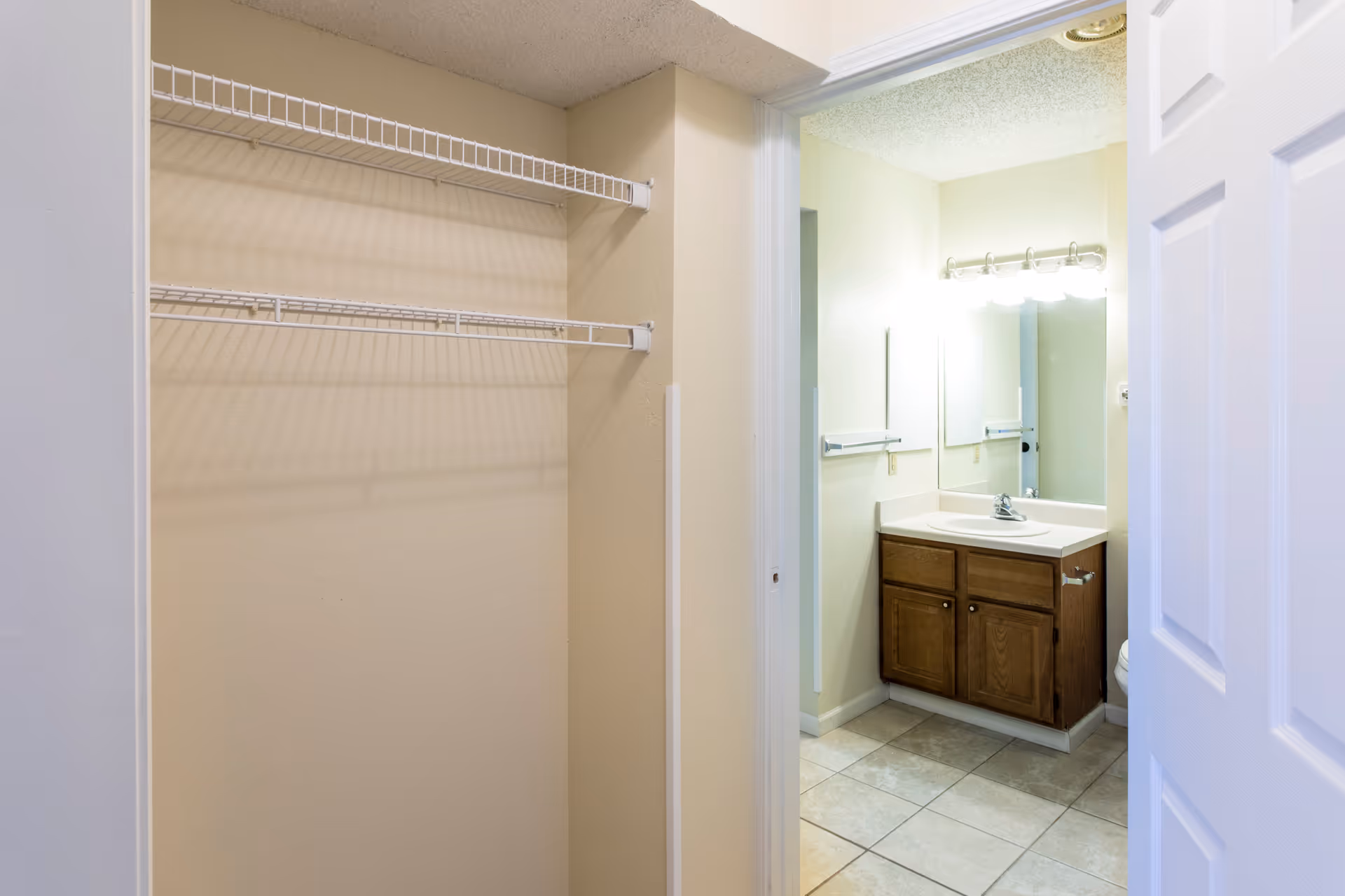 View of a small closet with two white wire shelves on the left and a bathroom on the right featuring a sink with a wooden cabinet, a large mirror, and bright vanity lights above it.
