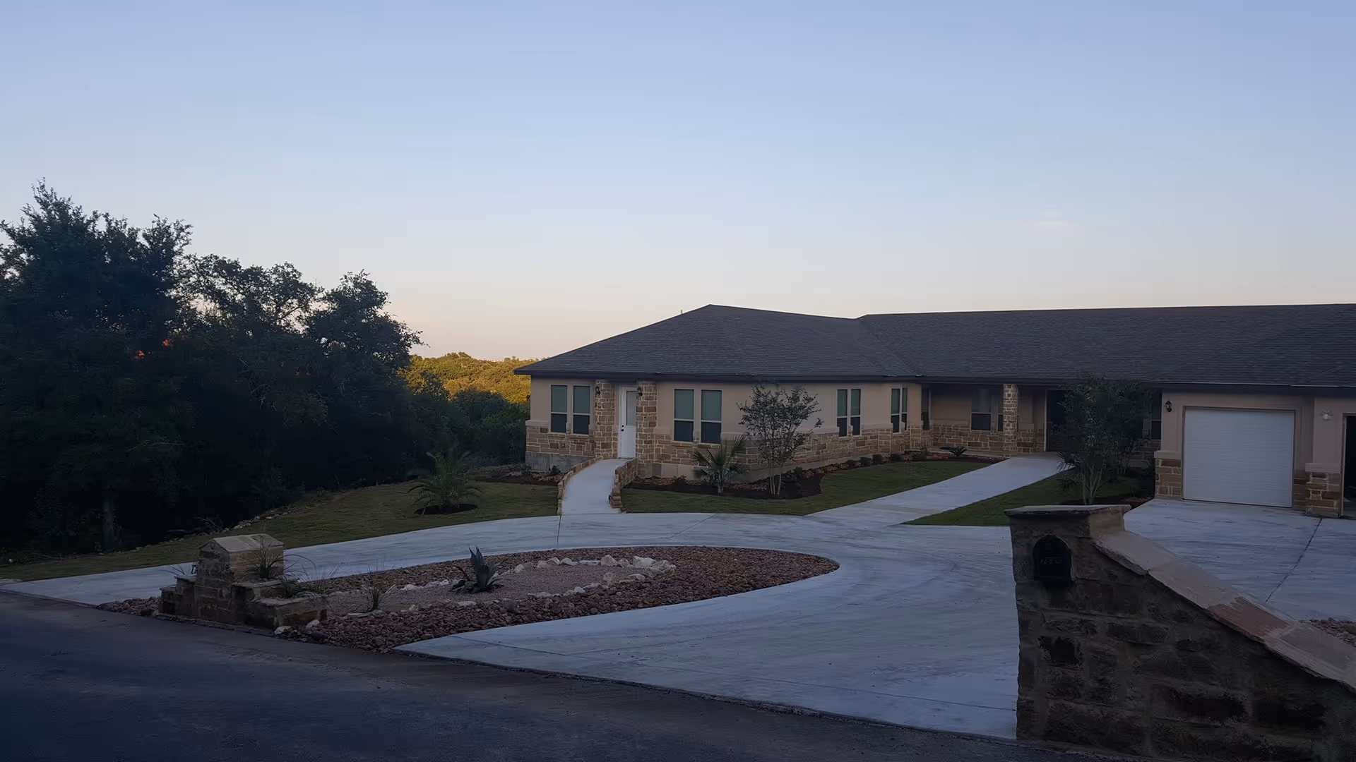 Exterior view of a single-story senior living facility building with stone and beige siding, a dark shingled roof, multiple windows, and a garage. The building is surrounded by a concrete driveway and landscaped areas with small trees and shrubs. Trees and greenery are visible in the background under a clear sky.