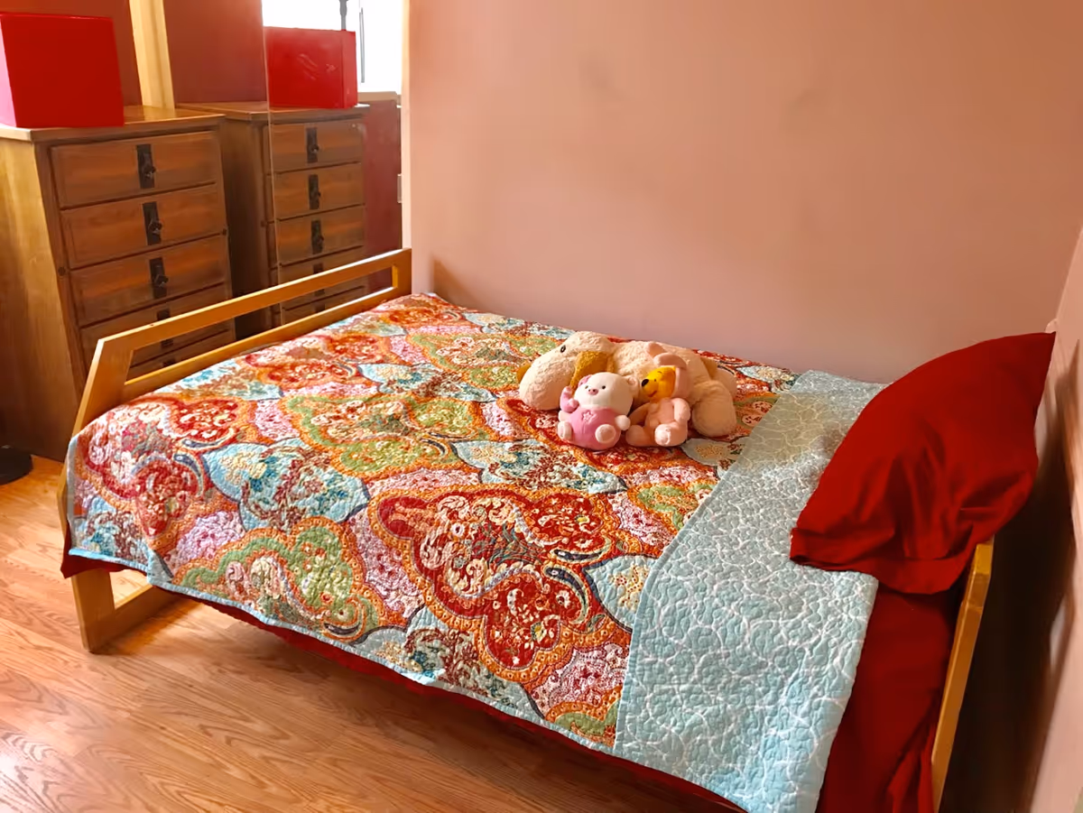 A bedroom with a wooden bed frame and a colorful quilt featuring red, orange, green, and blue patterns. There are three stuffed animals placed on the bed, and a red pillow rests against the wall. In the background, there is a wooden dresser with multiple drawers and a large mirror reflecting part of the room. The floor is wooden, and the walls are painted a soft pink color.