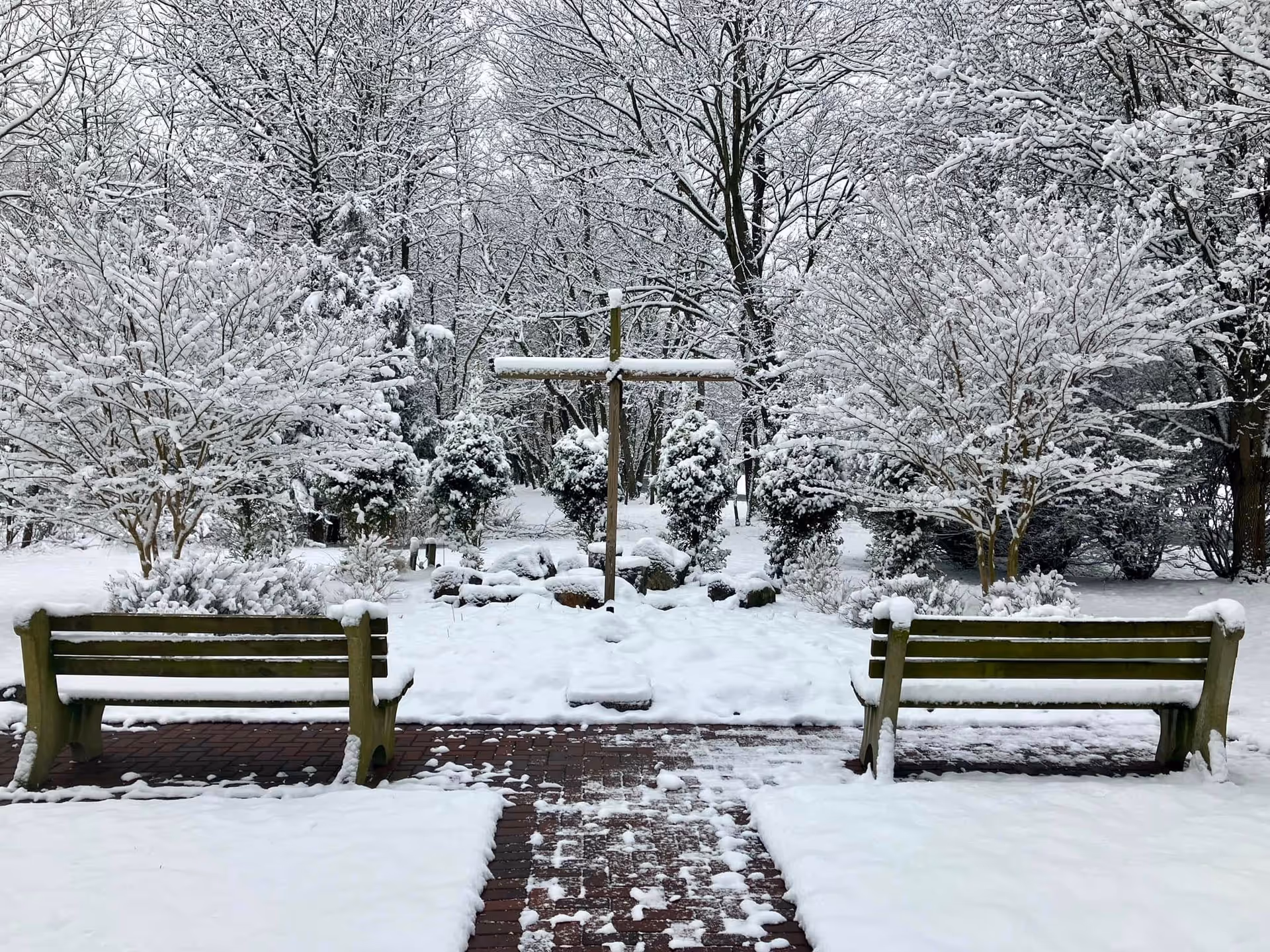 Snow-covered courtyard with two benches facing a wooden cross and trees blanketed in snow.