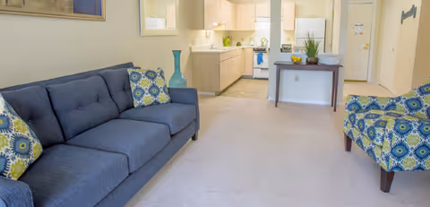 Bright furnished living room with a blue sofa, patterned armchair, and an open kitchen visible in the background.