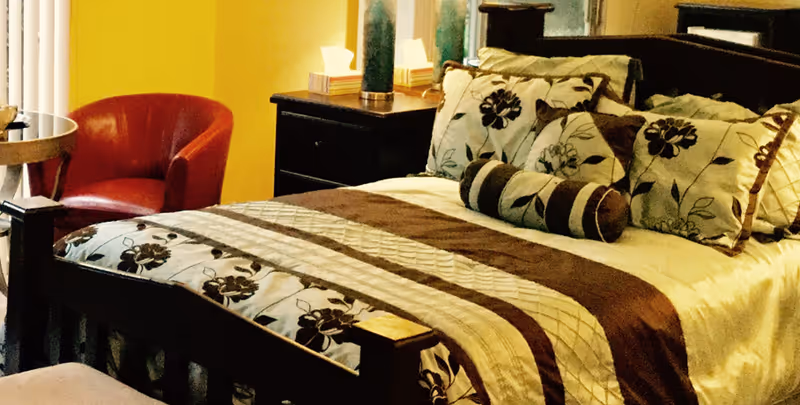 A cozy bedroom with a wooden bed frame, a bed made with floral patterned bedding and multiple pillows. There is a red armchair and a small round side table near a window with vertical blinds. A dark wooden nightstand with a lamp and some tissue boxes is next to the bed.