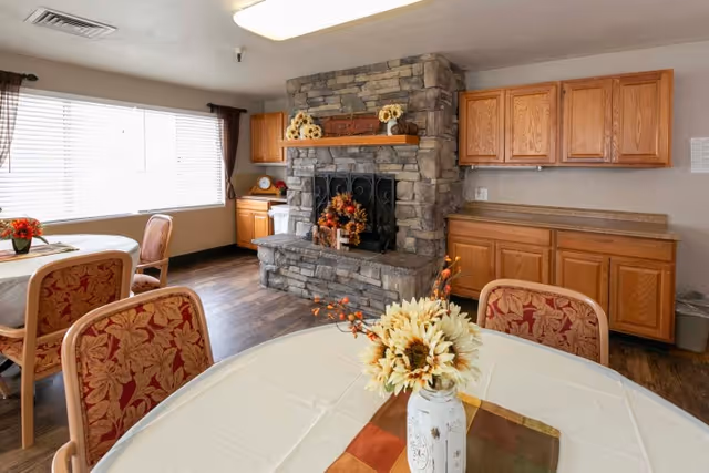 A dining area with round tables and floral centerpieces, upholstered chairs, a stone fireplace, and wooden cabinets.