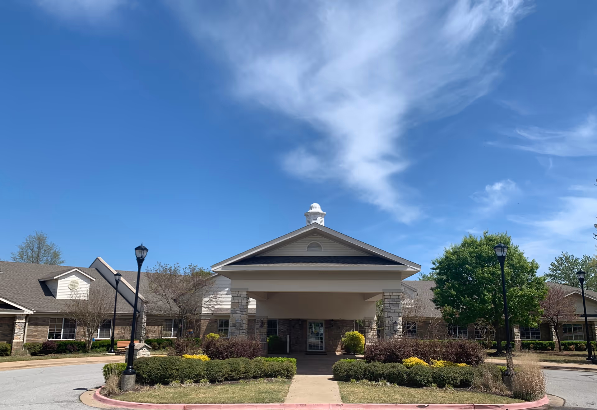 Front entrance of a single-story care facility with a covered porte-cochere, landscaped shrubs, lamp posts, and a blue sky.