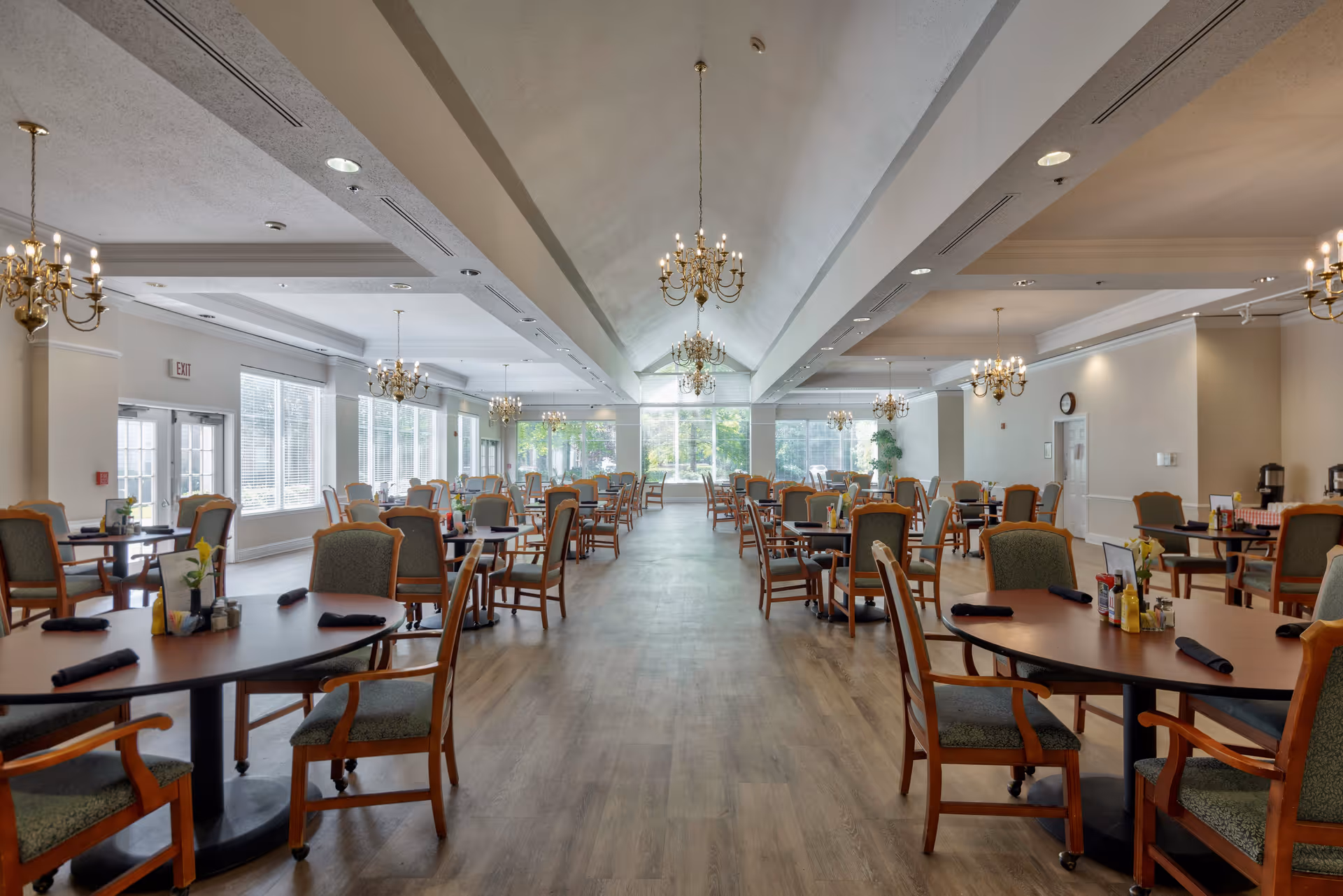 Spacious dining room with multiple round tables and wooden chairs arranged neatly. The room features large windows allowing natural light to fill the space, elegant chandeliers hanging from the ceiling, and a wooden floor. Each table is set with black napkins and condiments.