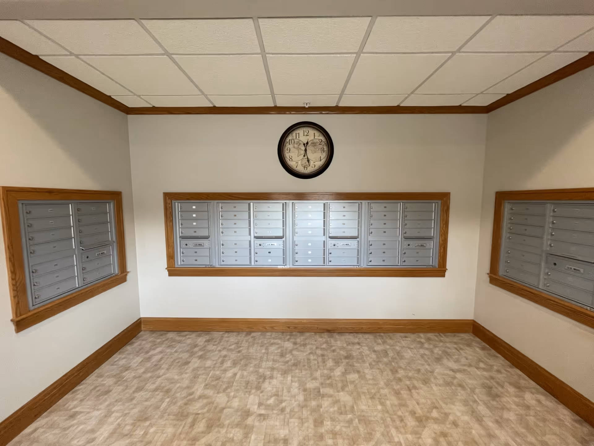 A room with beige walls and a tiled ceiling featuring a set of silver mailboxes mounted on the center wall, framed with wood trim. There are two mirrors on the left and right walls reflecting the mailboxes. A round clock is mounted above the mailboxes. The floor has a light brown patterned surface.