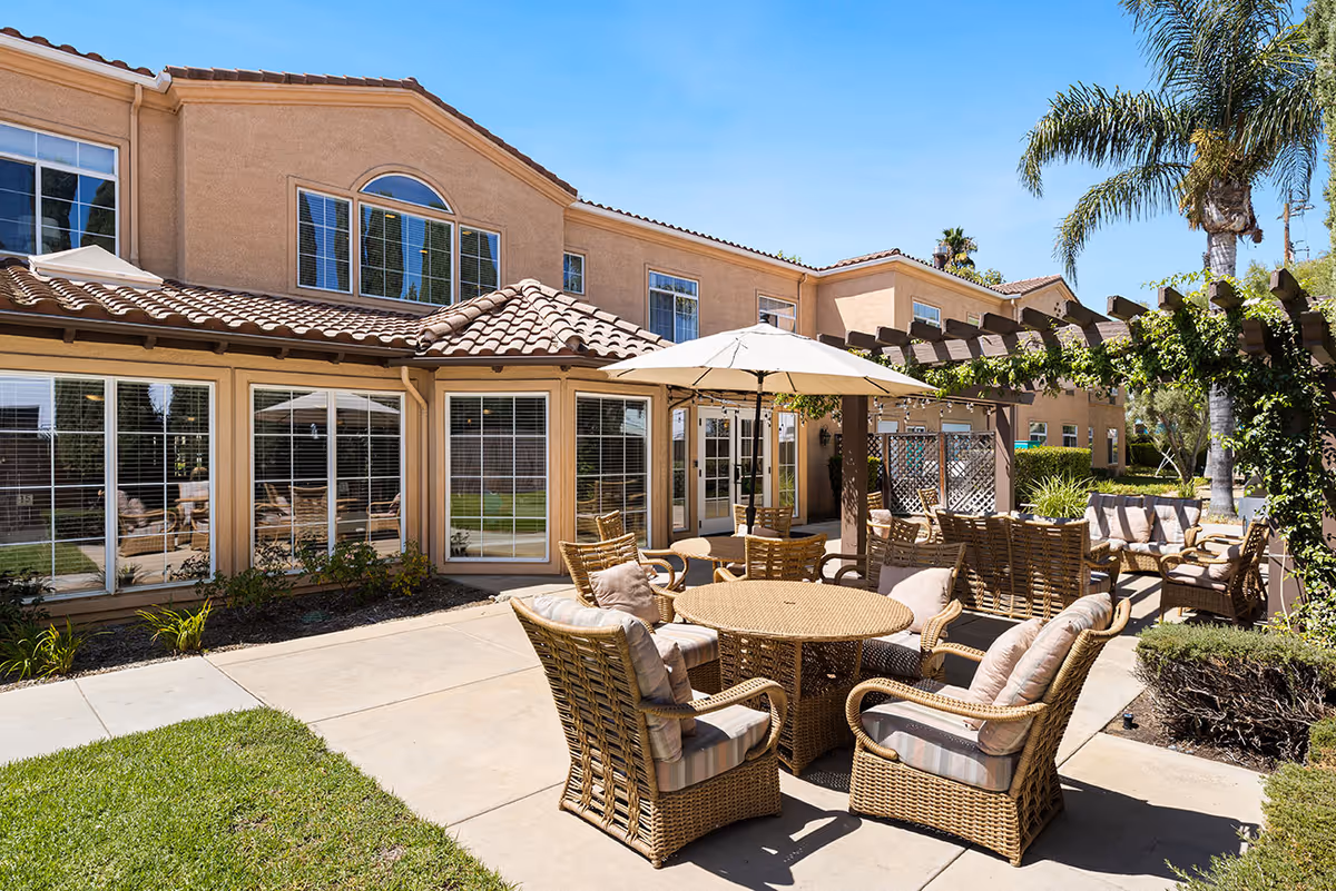 Sunny outdoor courtyard with wicker tables and chairs, umbrellas, a pergola, and a two-story stucco building in the background.