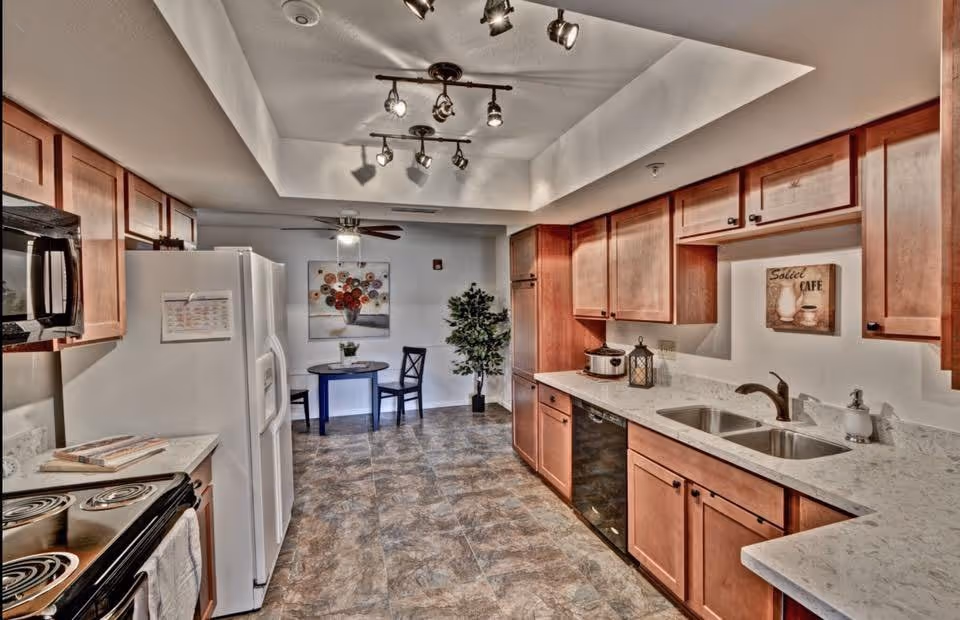 A clean and well-lit kitchen area with wooden cabinets, a white refrigerator, a black microwave, an electric stove, a double sink, and a dishwasher. There is a small round table with two chairs and a potted plant in the background. The floor is tiled, and the ceiling has track lighting and a ceiling fan.